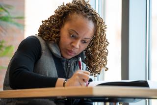 Brown skin woman with curly blondish brown locks holds a red pen while sitting at a table next to a large window