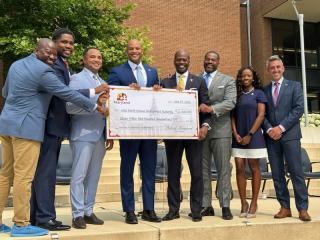 Eight people, seven men and a woman stand together holding an oversized check from the State of Maryland to the West North Avenue Development Authority