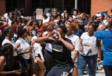 A student wearing a black Coppin State t-shirt cups his hands around his mouth and shouts enthusiastically while surrounded by a large, energetic crowd of students during an outdoor campus event.